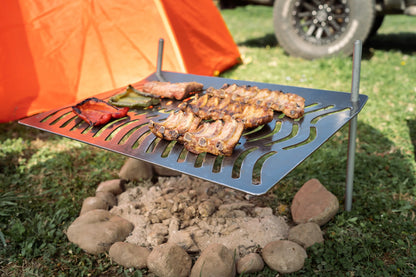 Humos Parrilla Camping grill with meat and vegetables cooking over an outdoor fire pit near an orange tent