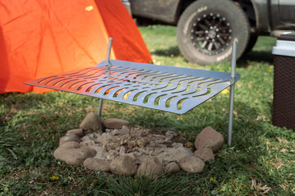 Humos Parrilla Camping metal grill setup over a stone fire pit with orange tent and vehicle in background
